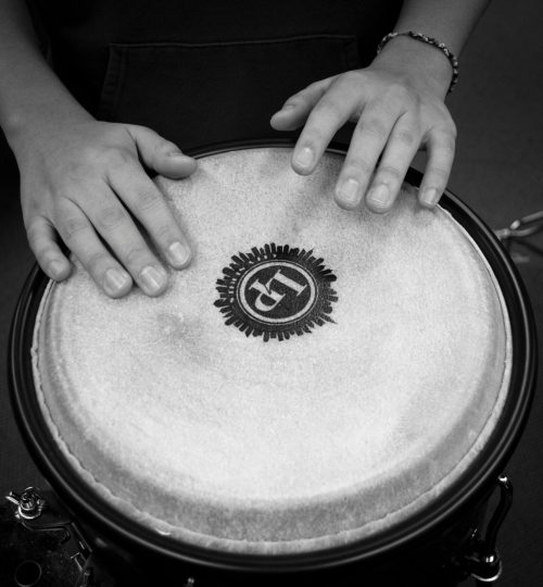 A black and white close-up shot of hands drumming on a bongo drum, emphasizing rhythm and music.